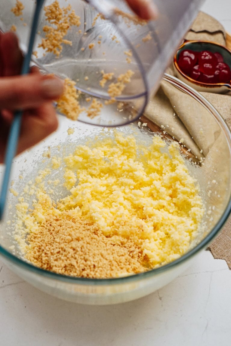 Hand adding crumbled mixture into a glass bowl with a yellow mixture, next to a bowl with red sauce and rolled dough on a beige cloth.