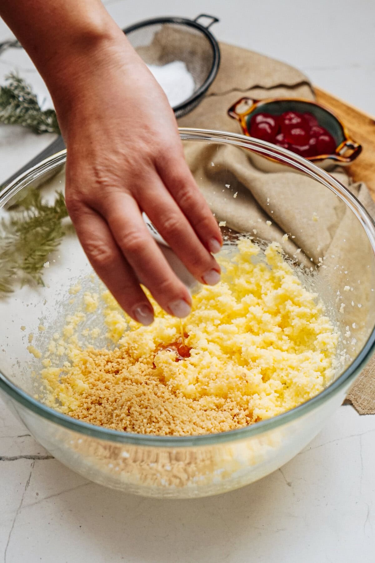 A hand adds liquid to a glass bowl containing a mixture of yellow and brown ingredients on a table, with a small dish of red jam nearby.