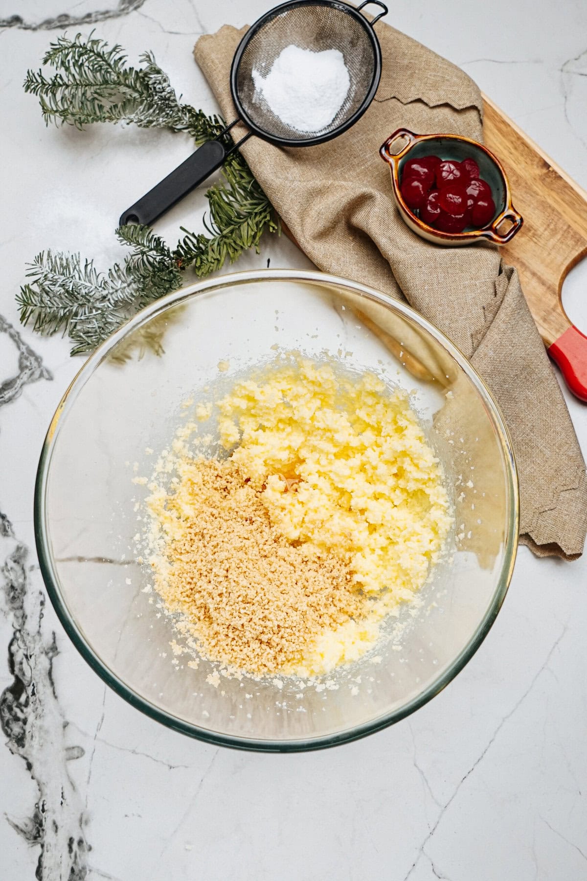 A glass bowl with mixed butter and sugar on a marble countertop. Nearby are a sieve with flour, a bowl of cherries, a wooden tray, a kitchen towel, and a sprig of greenery.