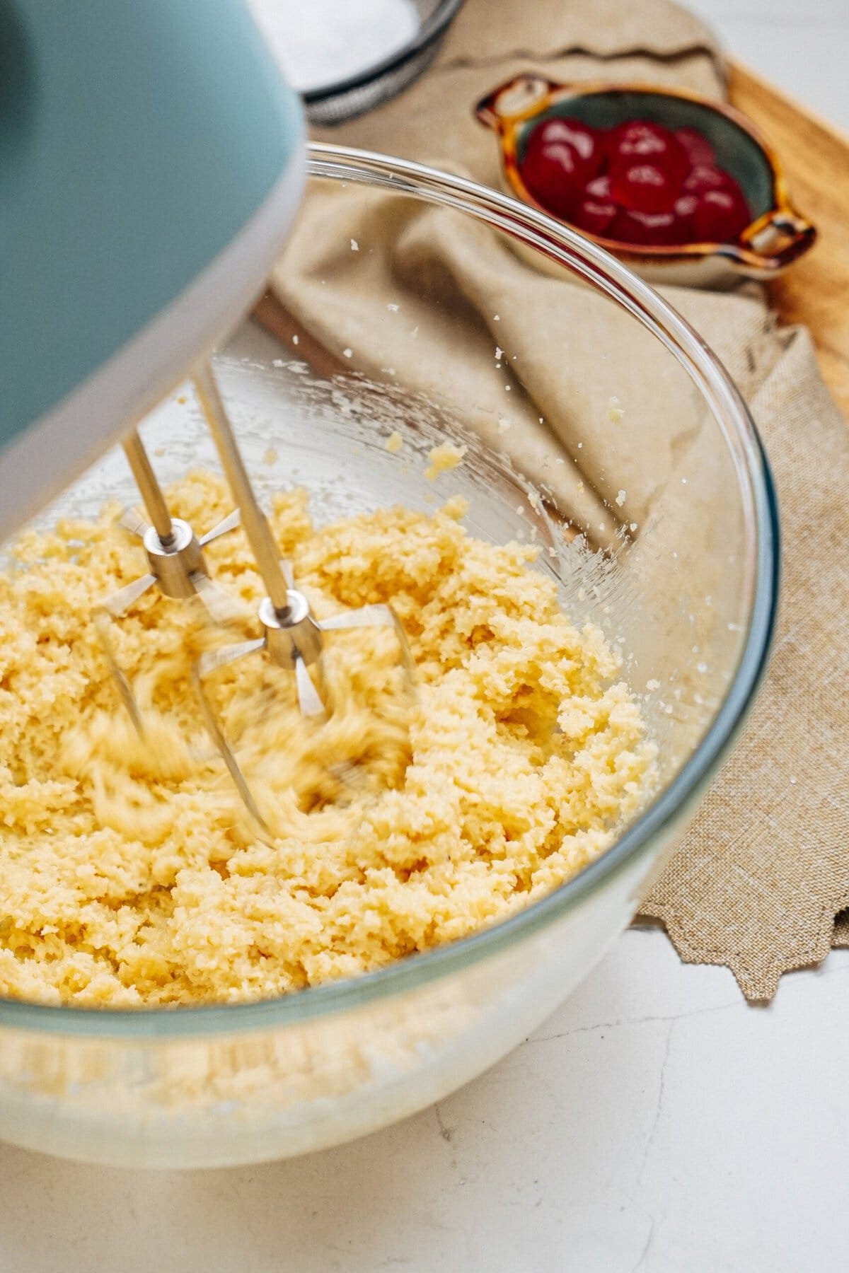 Electric mixer blending cake batter in a glass bowl, with a bowl of cherries and a cloth napkin in the background.