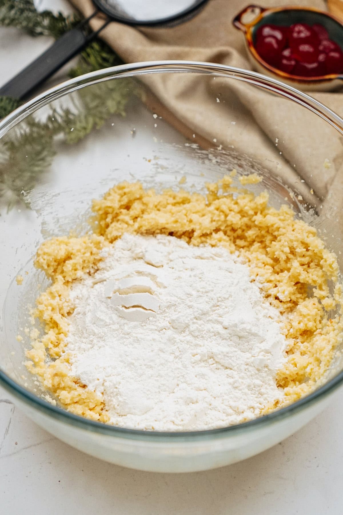 Glass bowl with flour and yellow dough mixture on a table. Nearby are a cloth, greenery, and a dish with red contents.