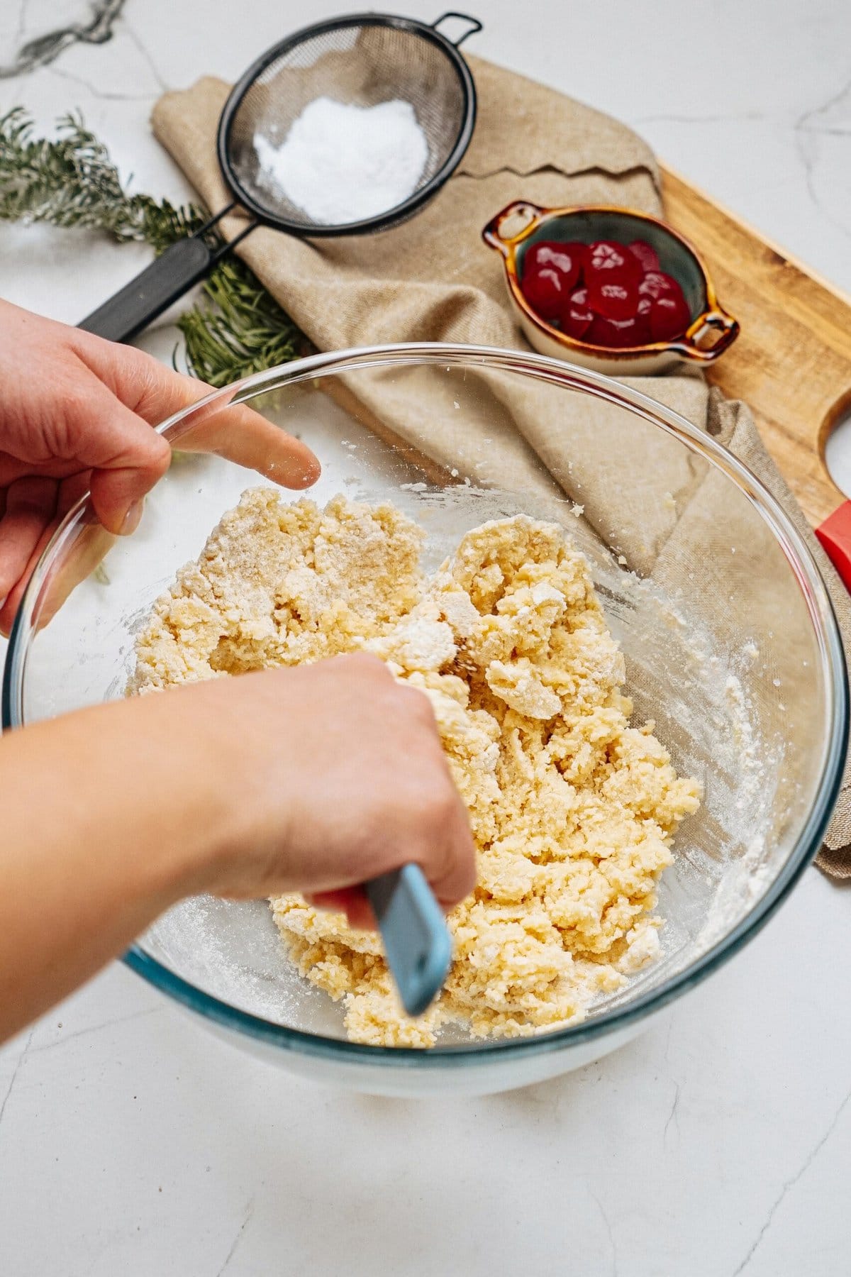 Hands mixing dough in a glass bowl. A sifter with flour, a dish with cherries, and a wooden board with a red knife are on the table.