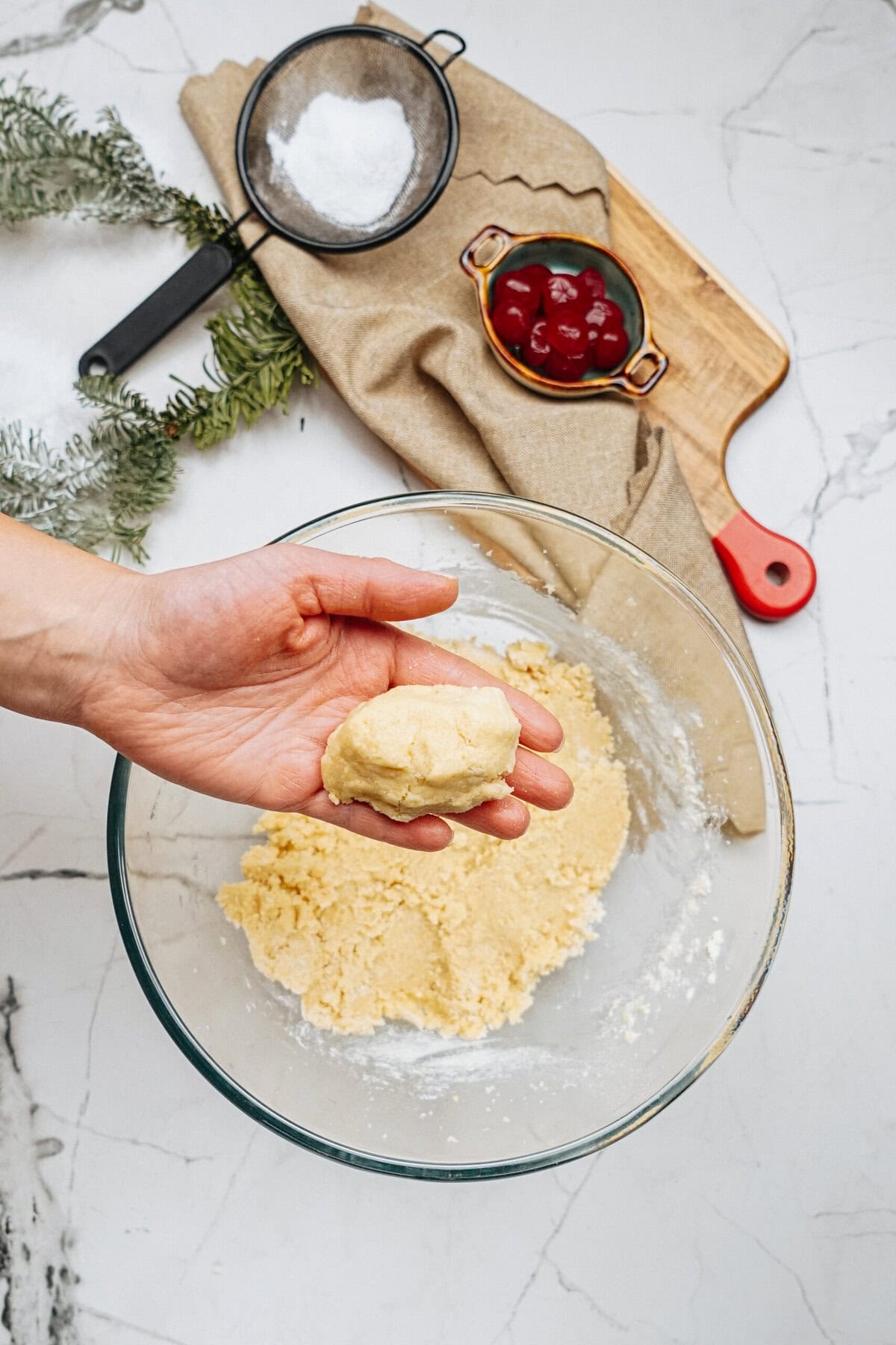 A hand holds a ball of dough over a glass bowl on a marble countertop. Nearby are a sifter with flour and a small bowl of cherries on a wooden board.