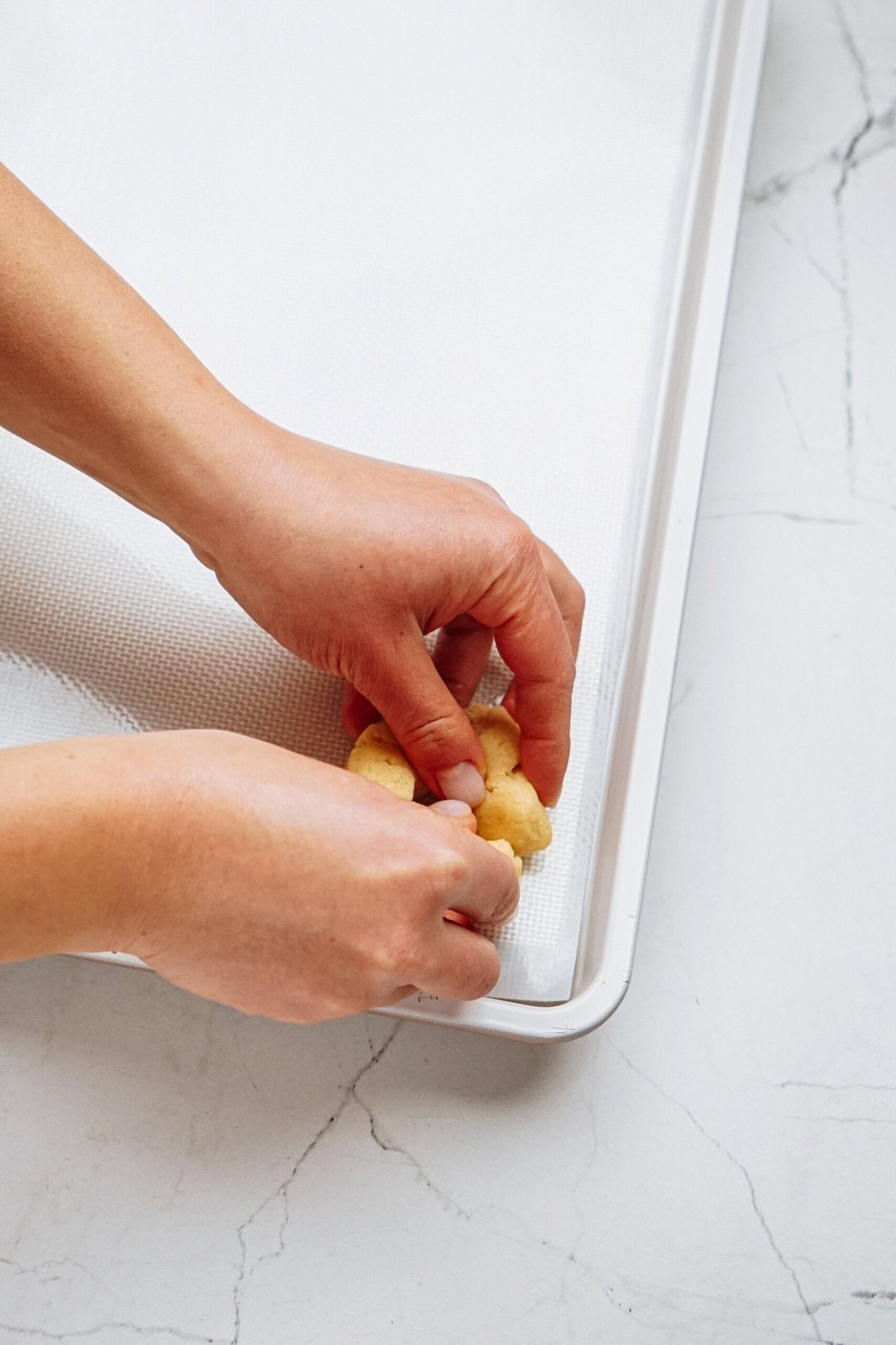 Hands pressing dough onto a baking sheet with a non-stick mat on a marble surface.