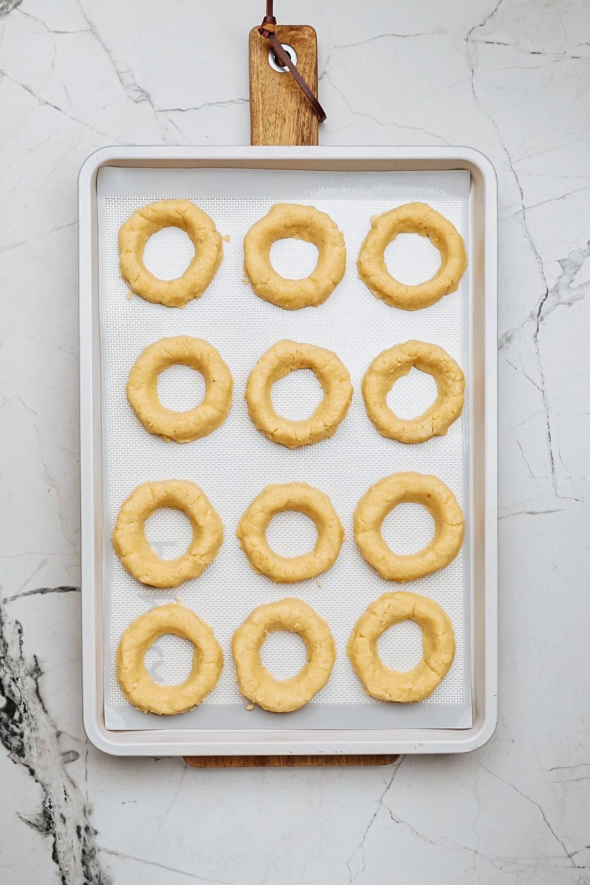 A baking sheet with 12 raw donut rings evenly spaced on parchment paper, placed on a marble surface.