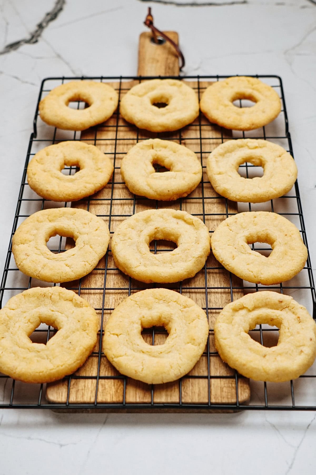 A dozen round cookies with holes in the center cooling on a wire rack placed on a wooden board.