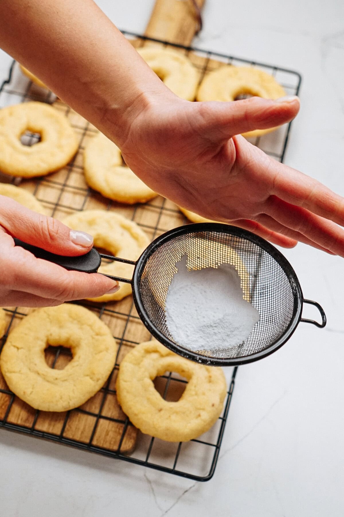 Hands sifting powdered sugar onto ring-shaped cookies on a cooling rack.