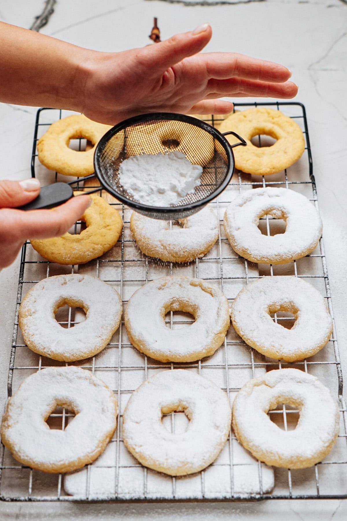 Hands sifting powdered sugar onto ring-shaped cookies on a cooling rack.