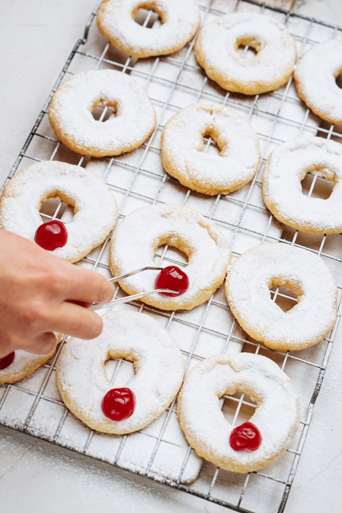 A hand places candied cherries on powdered sugar-covered ring-shaped cookies cooling on a wire rack.