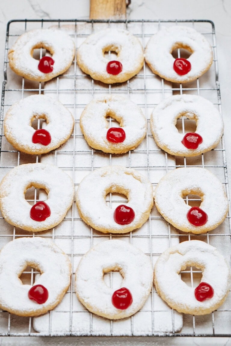 Round cookies dusted with powdered sugar and topped with red cherries are arranged in rows on a cooling rack.