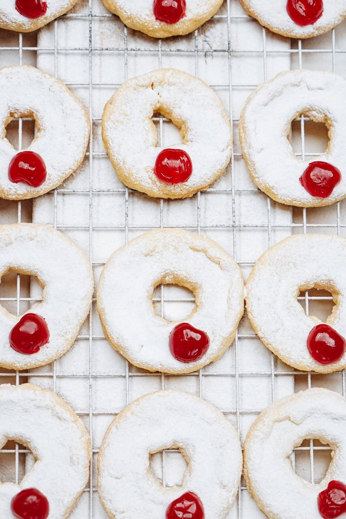 Round cookies with white icing and a red cherry on top are arranged on a cooling rack.