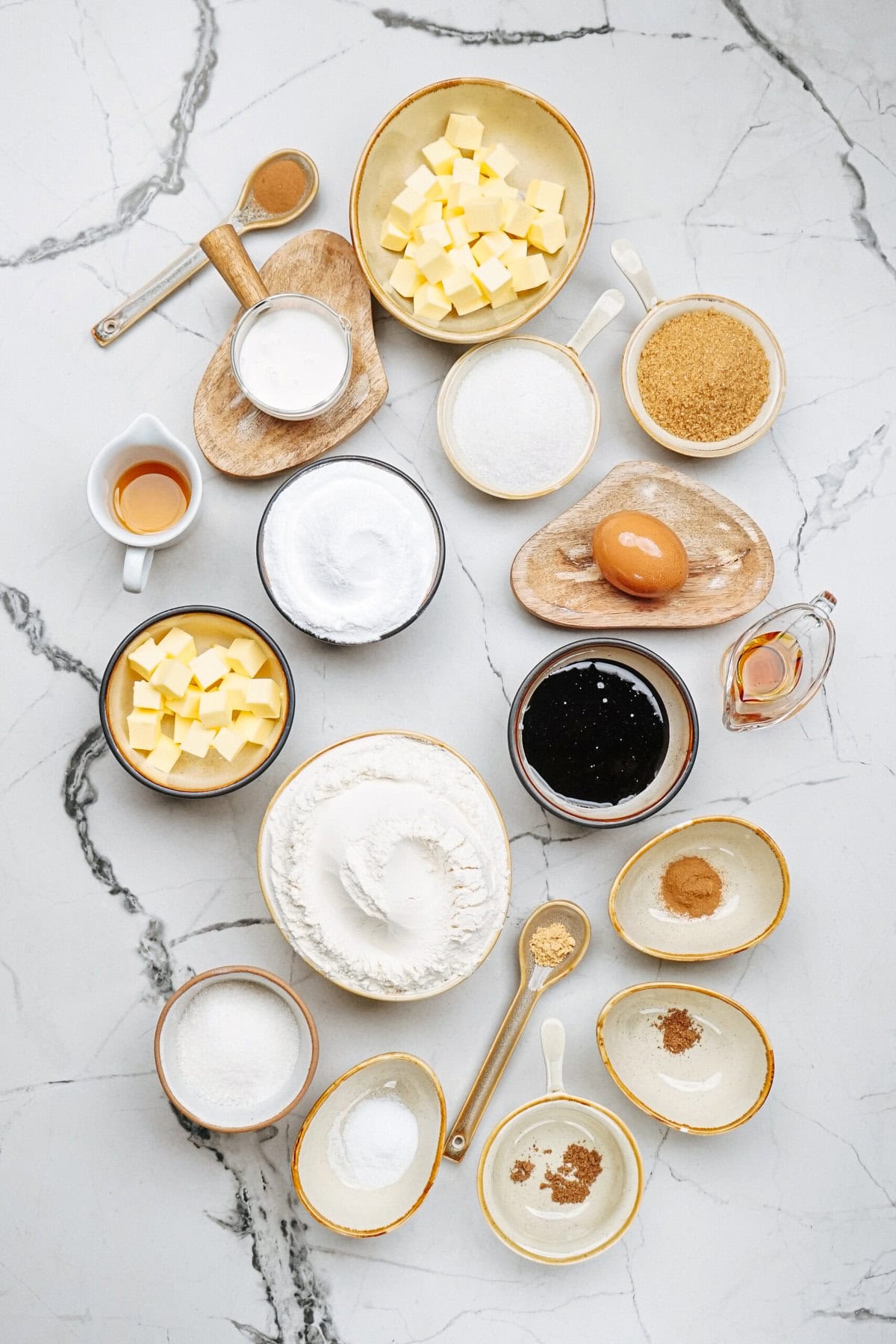 A variety of baking ingredients, perfect for gingerdoodle cookies, are arranged neatly on a marble surface: butter, sugar, flour, an egg, milk, vanilla extract, baking powder, and spices.