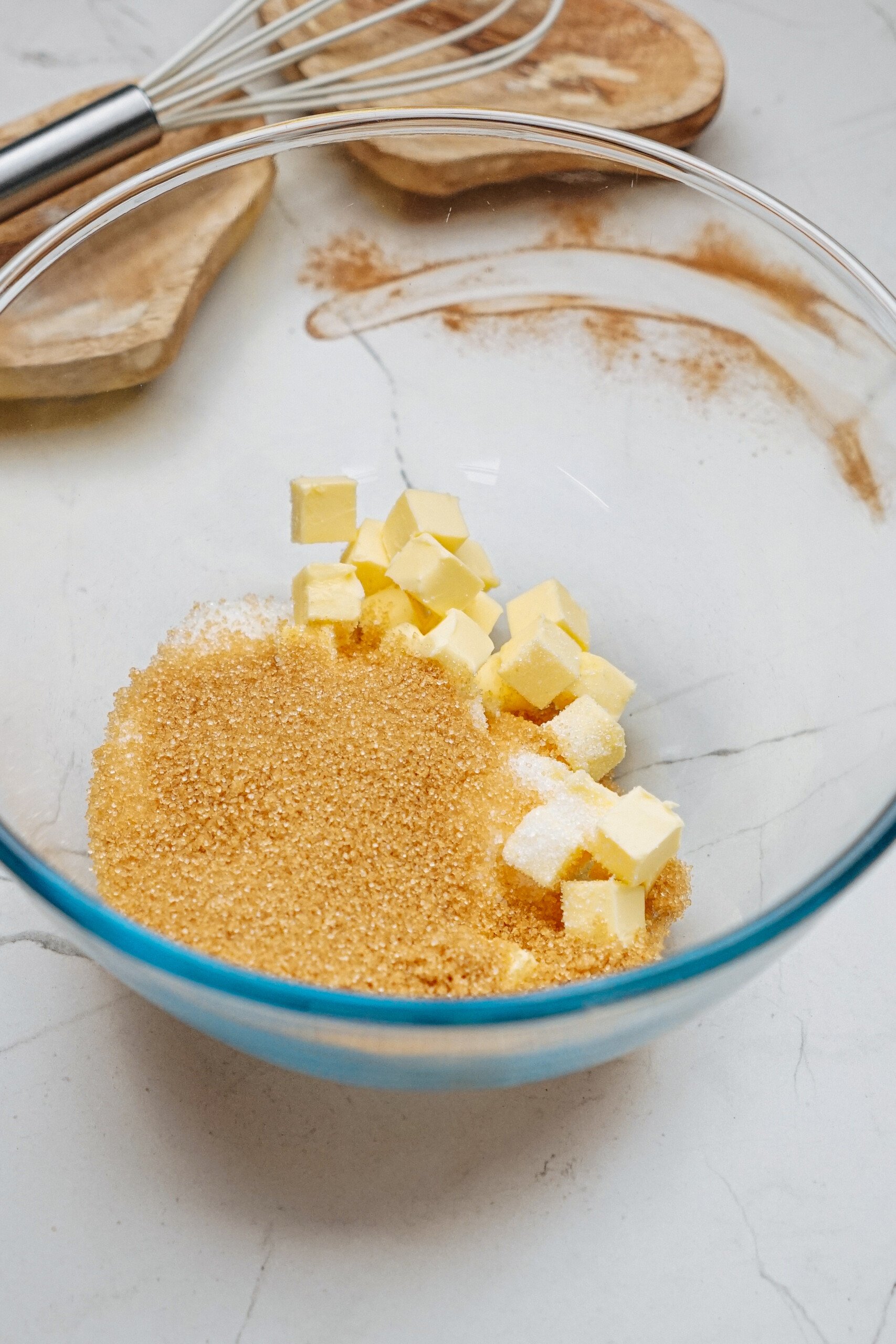 Cubed butter and brown sugar in a glass mixing bowl on a marble countertop, poised for the creation of gingerdoodle cookies, with a whisk and wooden board subtly gracing the background.