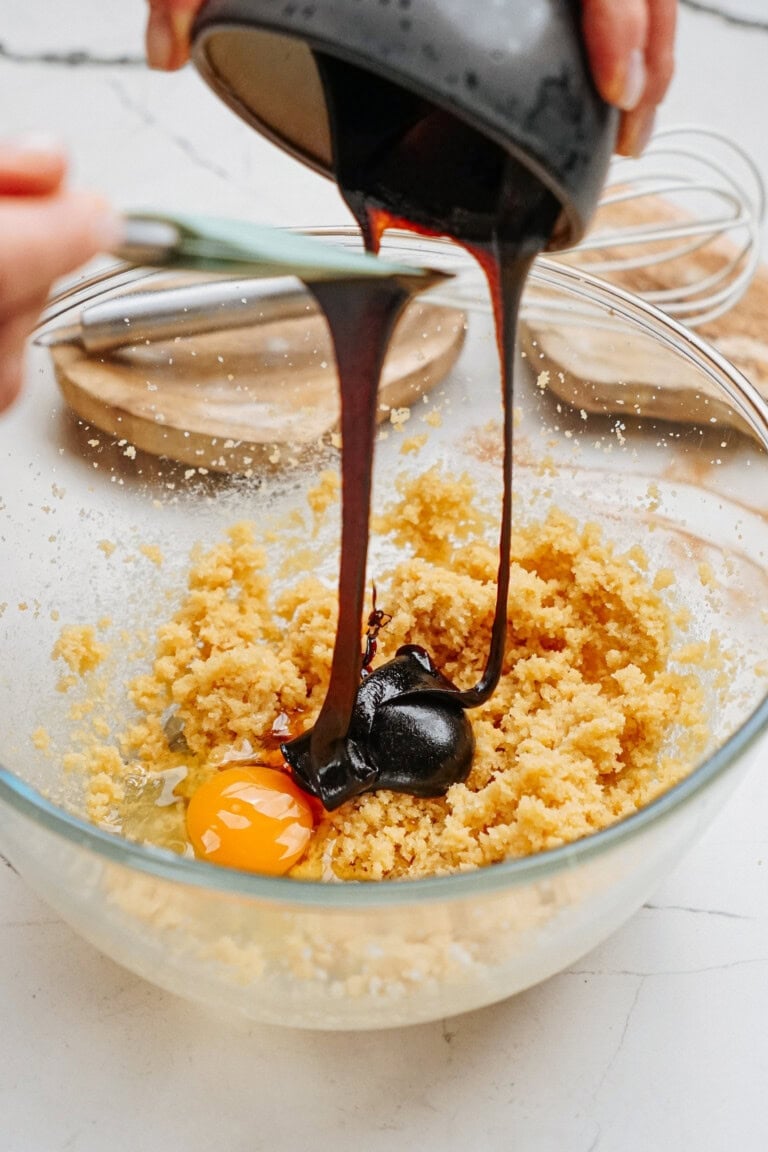 Molasses being poured into a glass bowl with ginger cookie dough and egg on a kitchen counter.