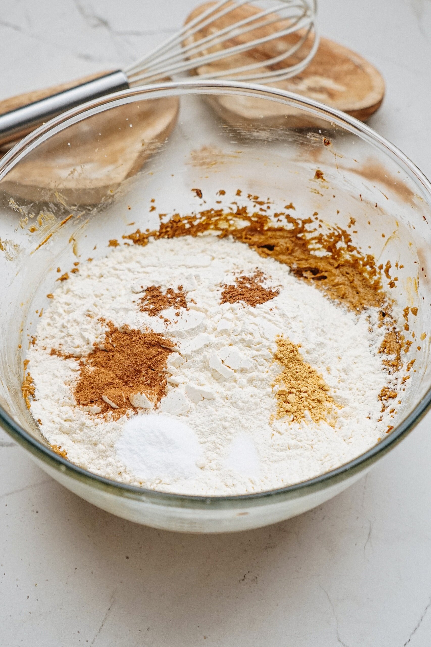 A clear glass bowl contains flour, baking soda, cinnamon, and other powdered ingredients needed for gingerdoodle sandwich cookies, with a whisk nearby on a marble countertop.