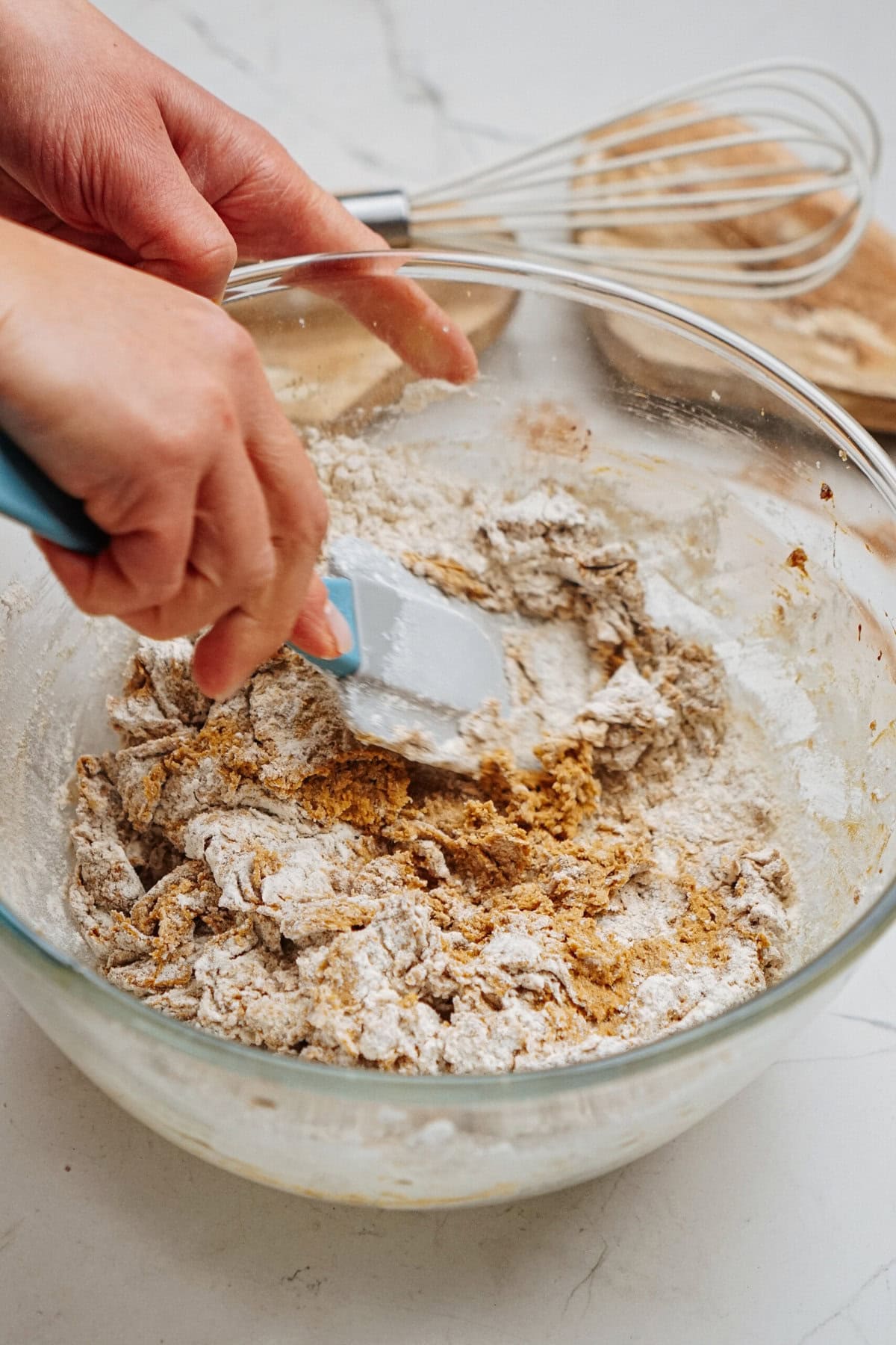 Hands mixing dry ingredients in a glass bowl with a blue spatula, preparing gingerdoodle sandwich cookies, next to a whisk and a wooden board.