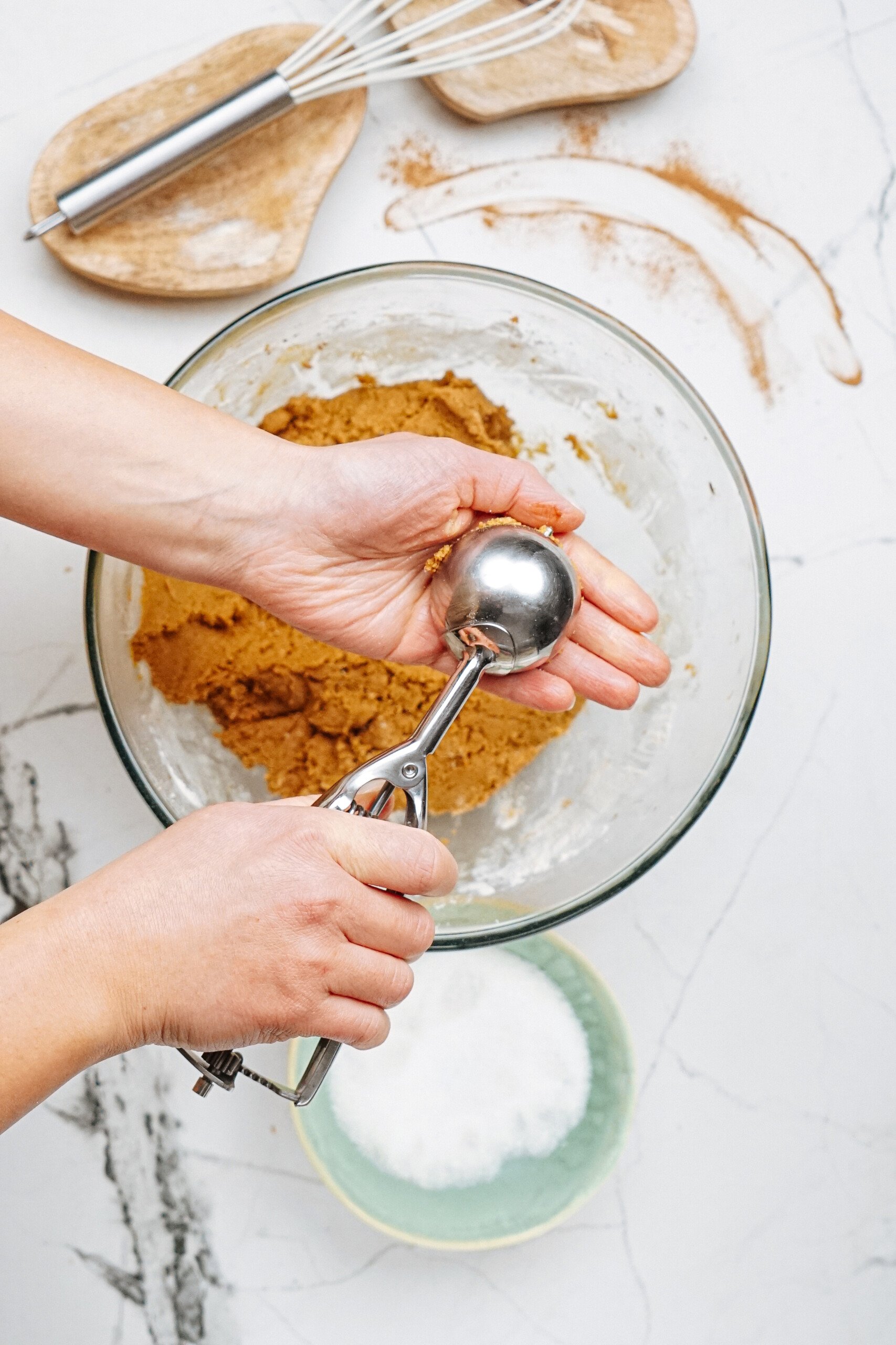 A person uses a metal scoop to portion dough from a glass bowl on a marble countertop. Nearby, a bowl of sugar and a whisk hint at more delicious creations to come.