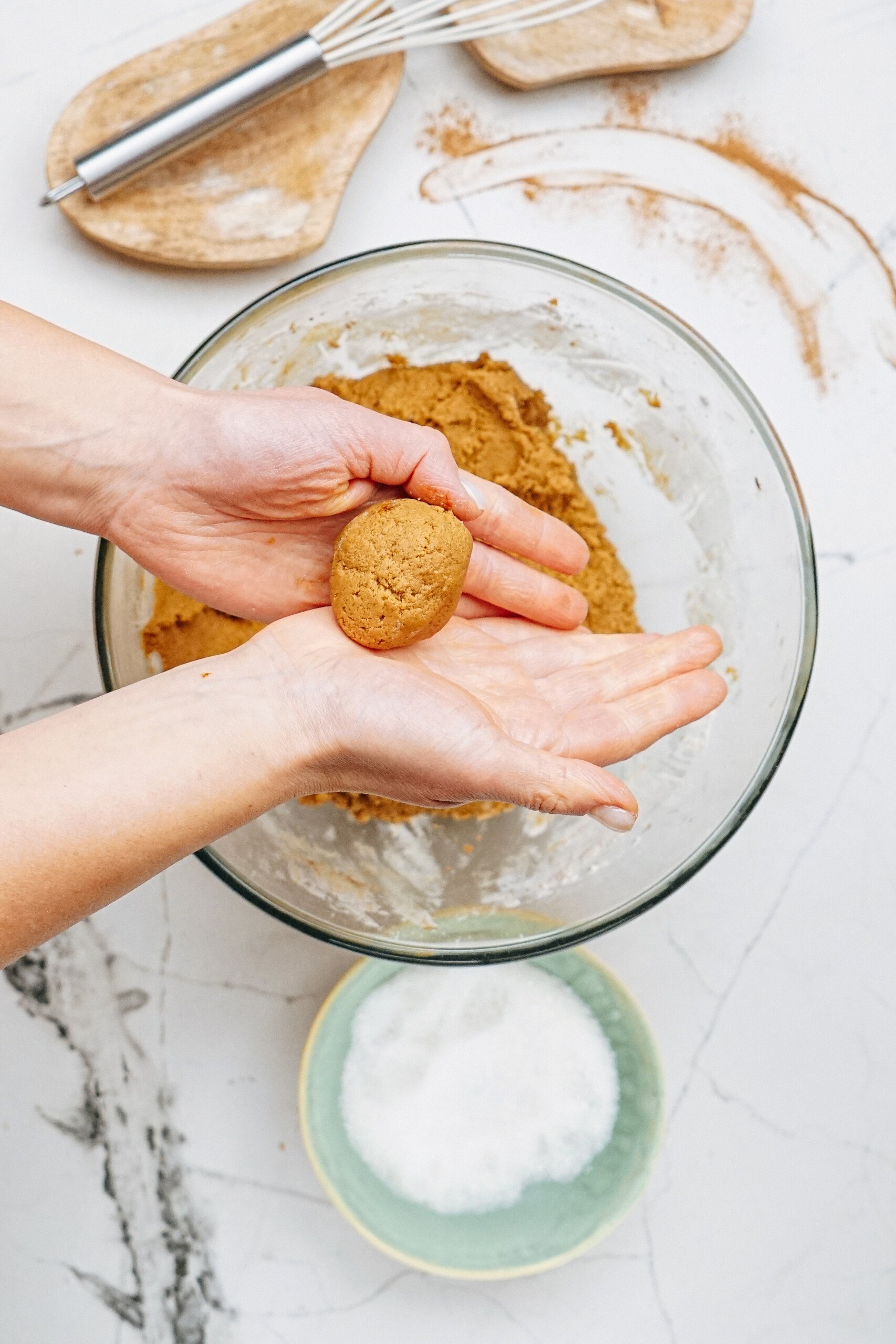 Hands shaping cookie dough over a glass bowl, with a small bowl of sugar nearby on the marble countertop. A whisk is in the background.