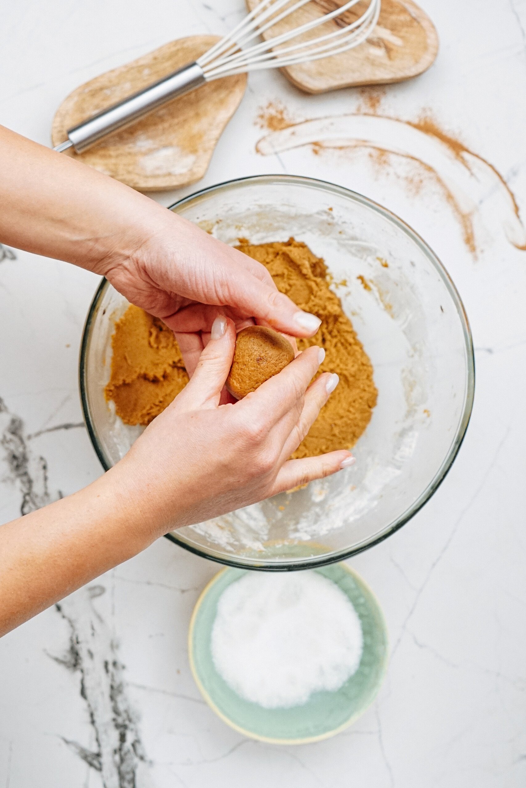 Hands shaping cookie dough over a bowl, with a whisk and small bowl of sugar nearby on a marble counter.