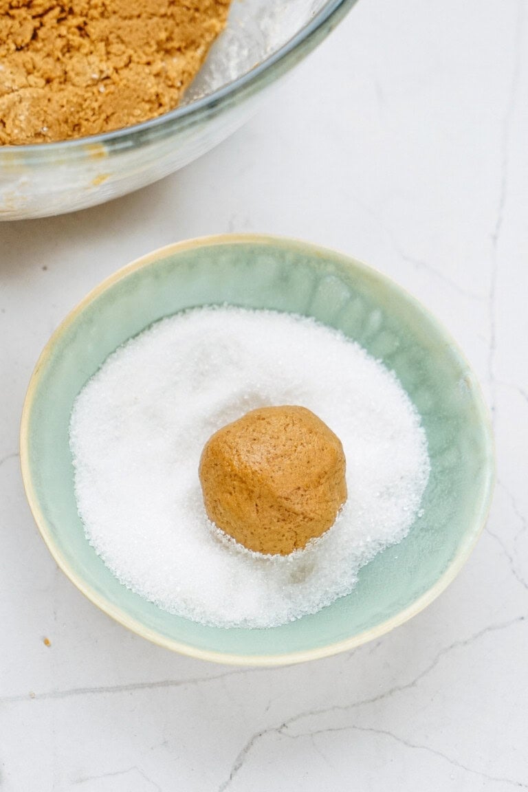 A ball of cookie dough sits in a bowl of white sugar, ready for coating. A larger bowl with more dough waits eagerly in the background on a pristine white countertop.