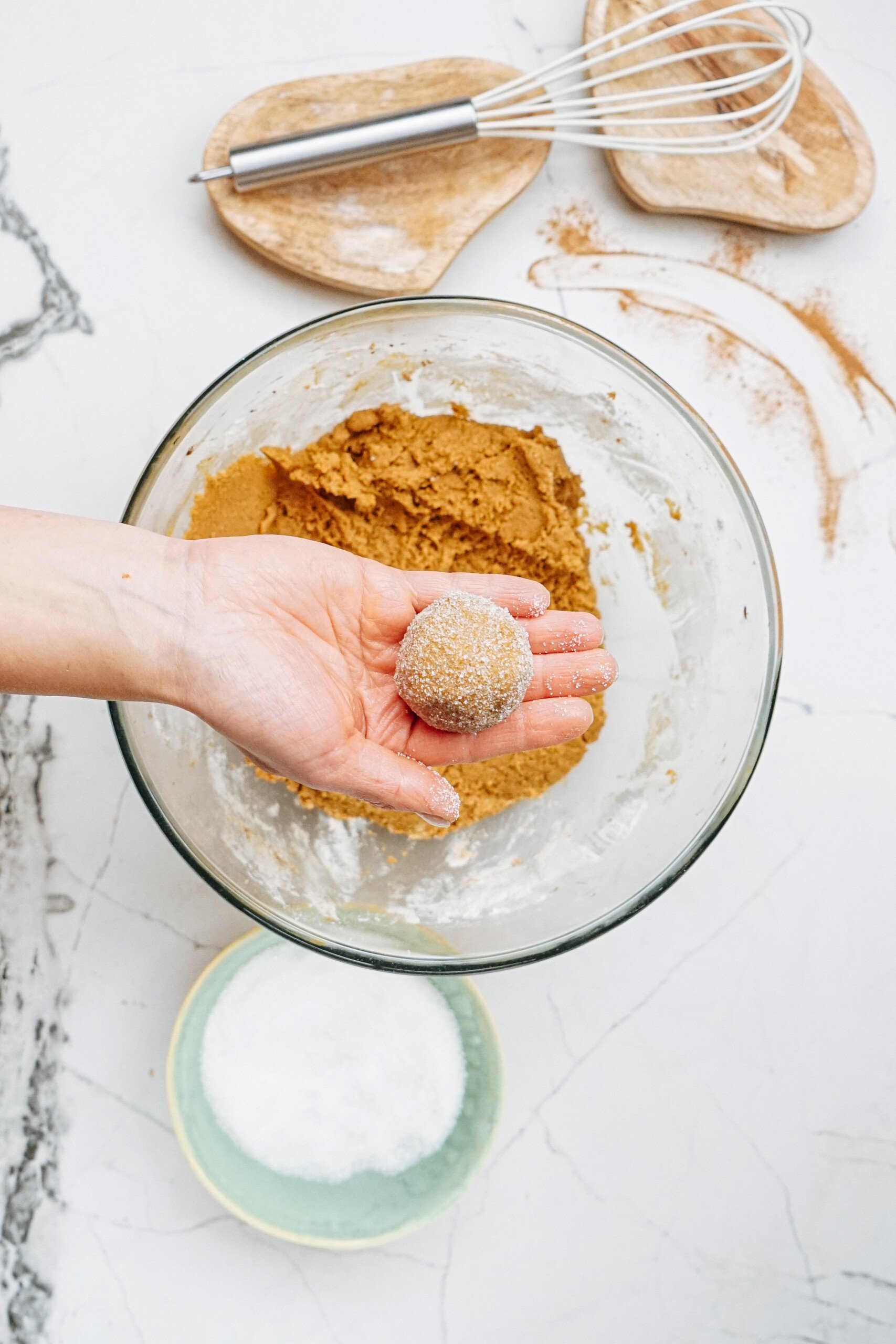 A hand holds a sugar-coated dough ball over a mixing bowl filled with cookie dough, ready to be transformed into gingerdoodles. Nearby, a whisk and two wooden boards rest on the marble surface alongside a bowl of sugar.