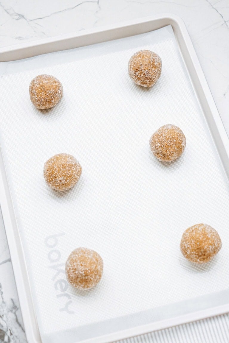 Six sugar-coated dough balls are evenly spaced on a white baking tray lined with a non-stick sheet, exuding the rich aroma of hazelnut shortbread.