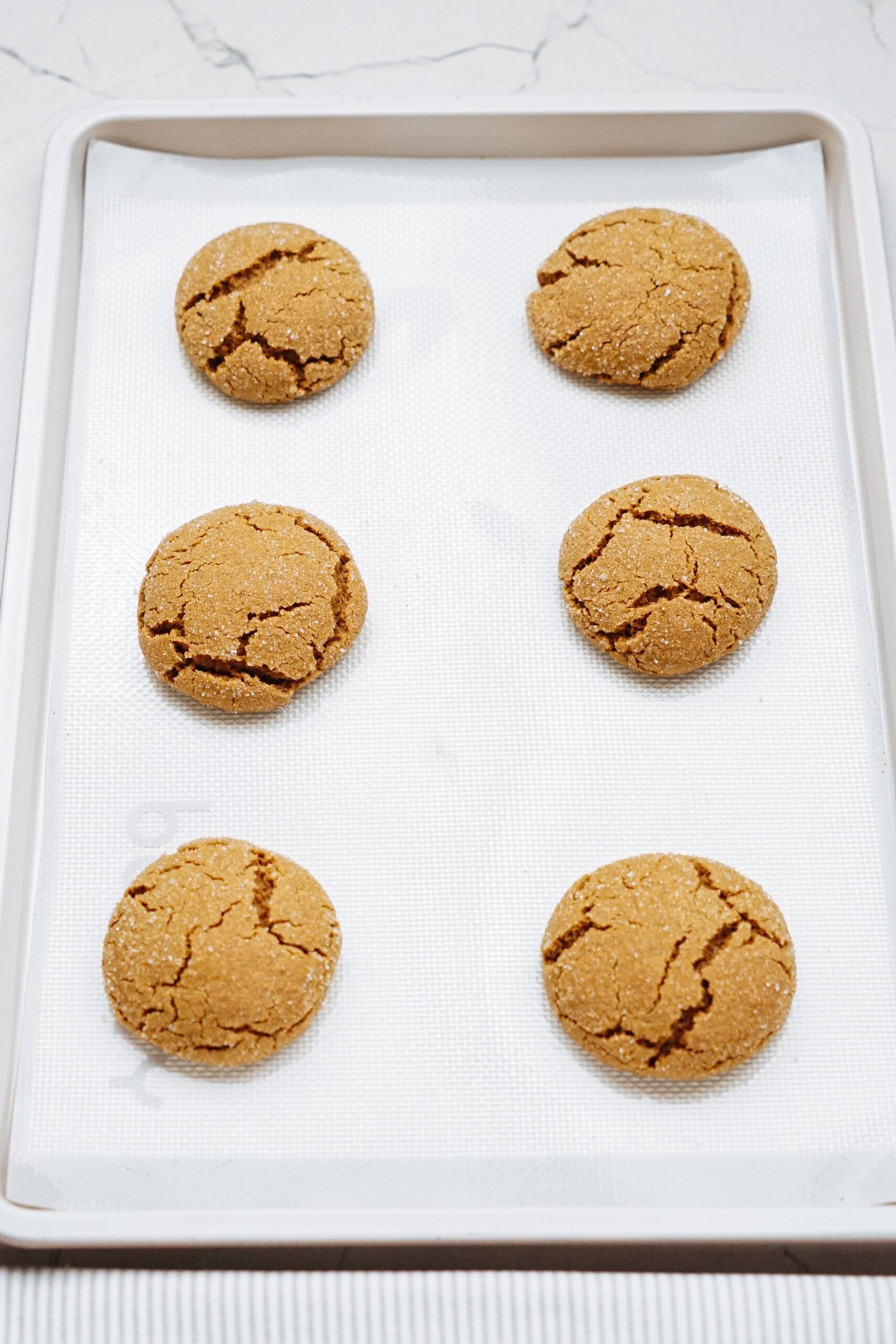 Six round cookies rest on a baking sheet lined with parchment paper, placed on a light surface.