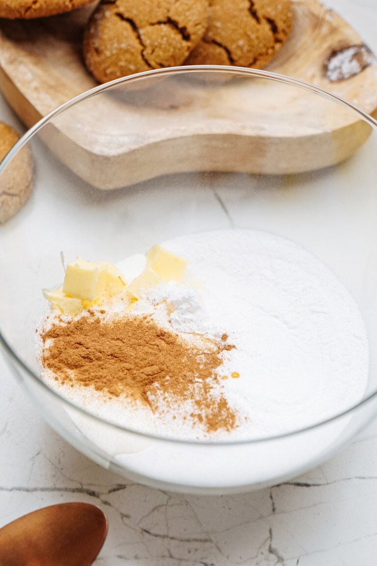 A glass bowl with flour, sugar, cinnamon, butter cubes, and baking powder sits on a marble surface surrounded by baking utensils and hazelnut shortbread cookies.