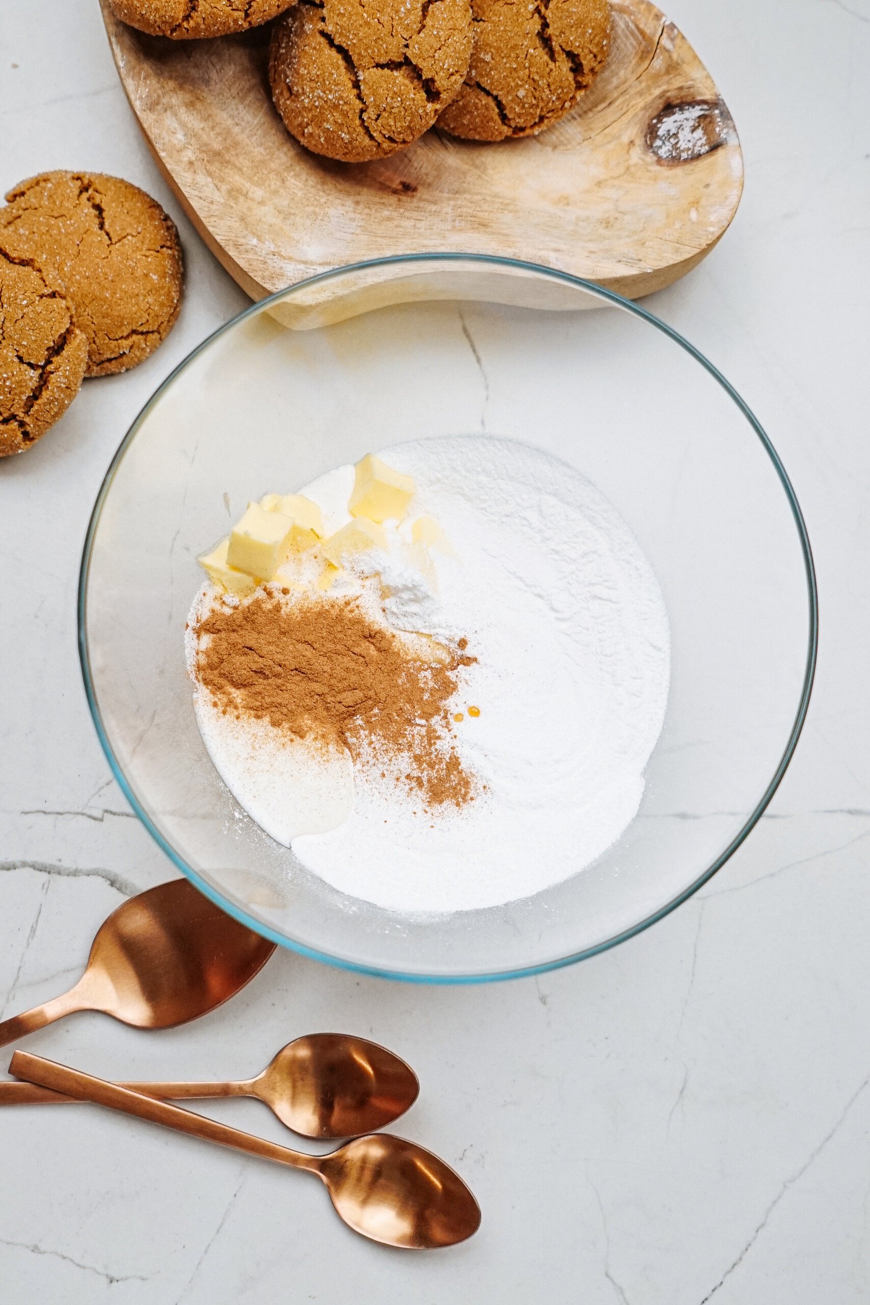 A glass bowl with flour, sugar, cinnamon, and butter sits on a marble surface. Three copper spoons rest nearby, while hazelnut shortbread cookies tempt from a wooden plate in the background.