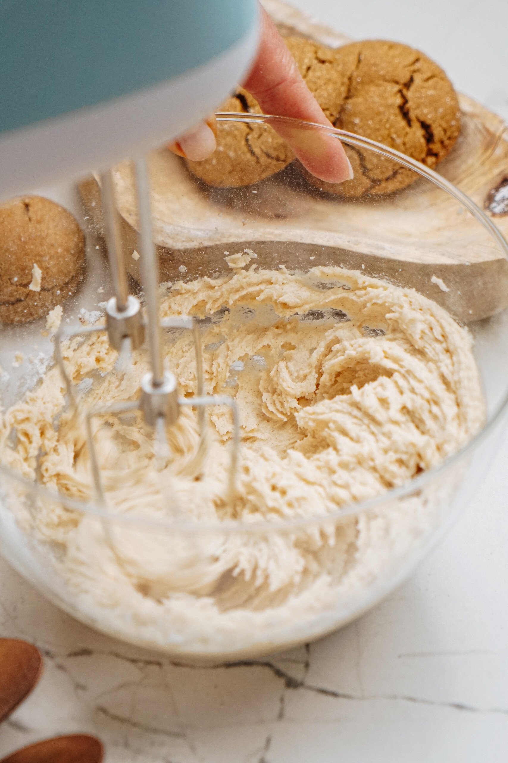 A hand mixing hazelnut shortbread cookie dough in a glass bowl with an electric mixer, next to a wooden board with freshly baked cookies.