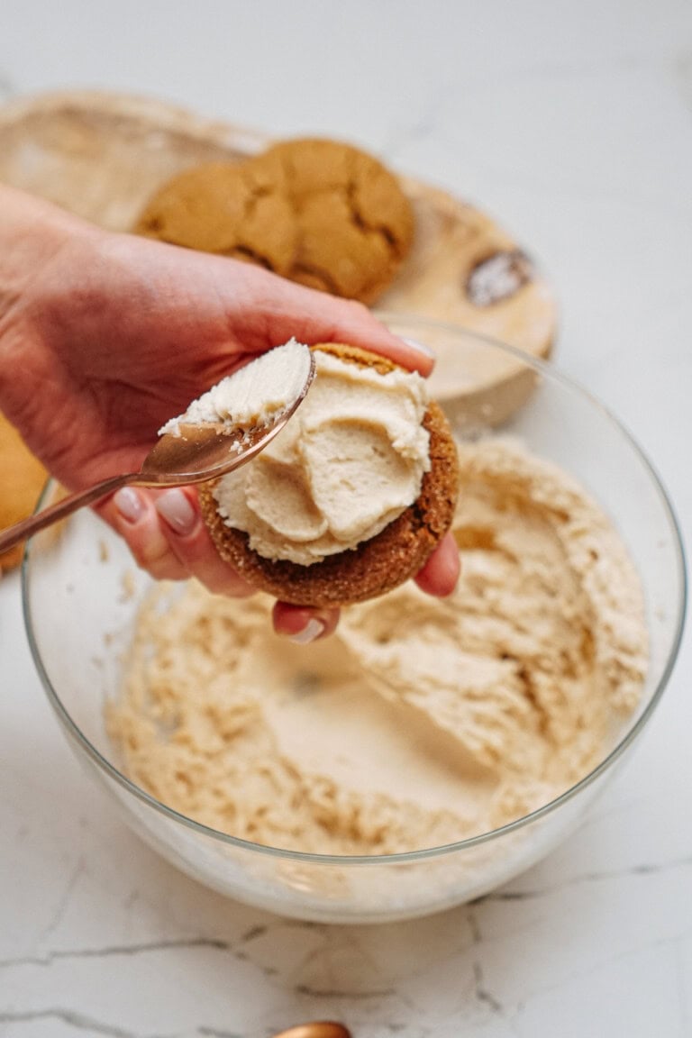 A hand gently spreads cream onto a Hazelnut Shortbread cookie with a spoon, while a bowl filled with creamy goodness and more cookies waits invitingly in the background.