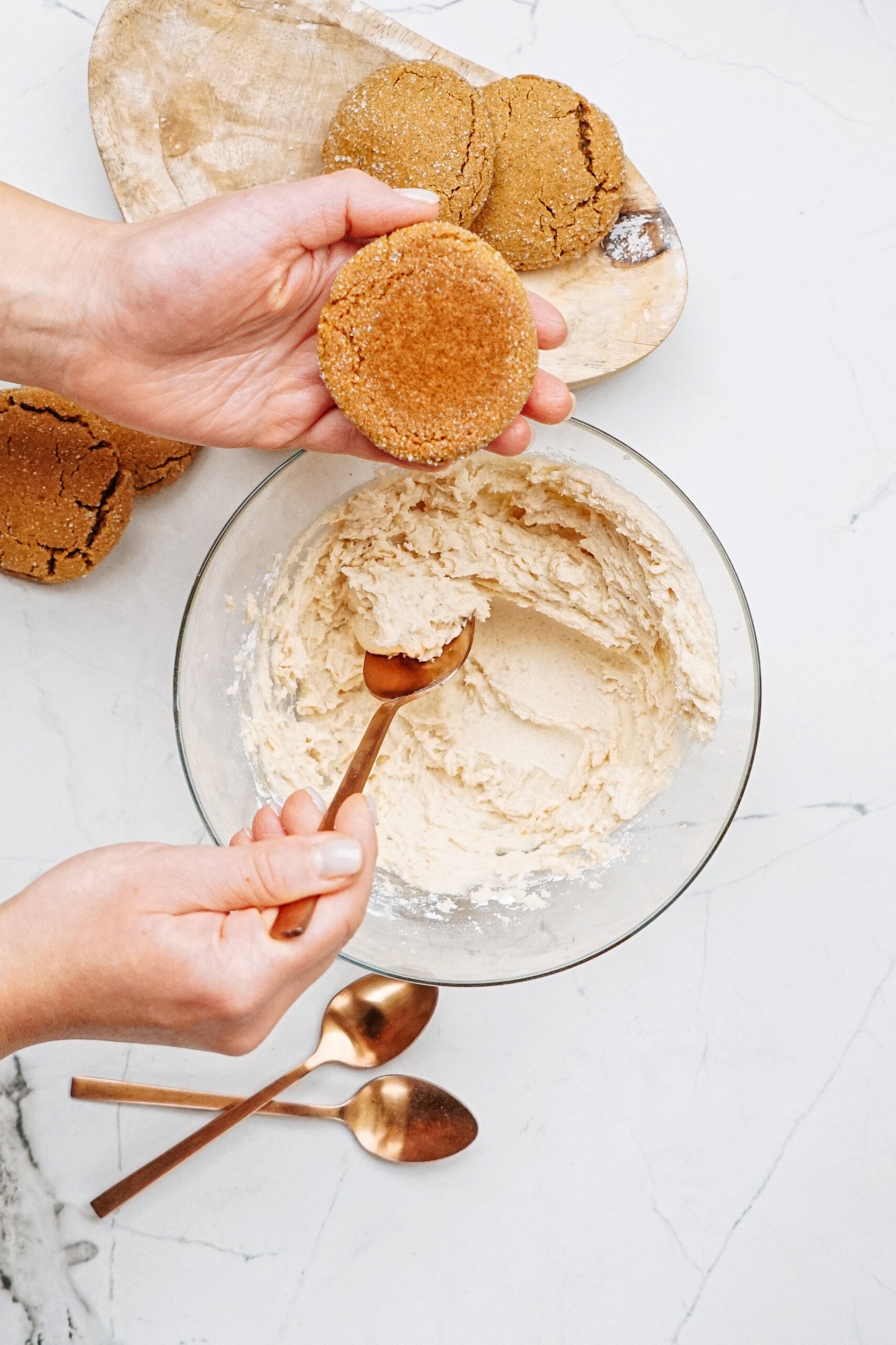 Hands holding a hazelnut shortbread cookie, while a spoon mixes dough in a bowl on the marble surface. Nearby, cookies rest on a wooden tray with two bronze spoons glinting softly beside them.