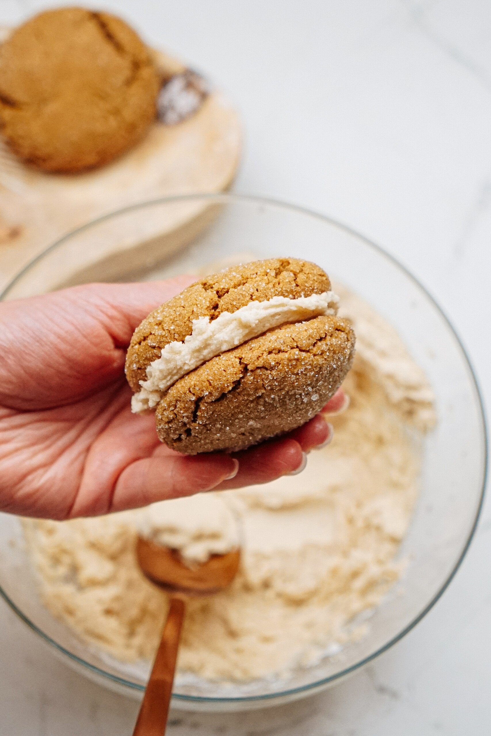 A hand holds a ginger cookie sandwich with cream filling over a bowl of flour and a wooden spoon, reminiscent of crafting hazelnut shortbread.