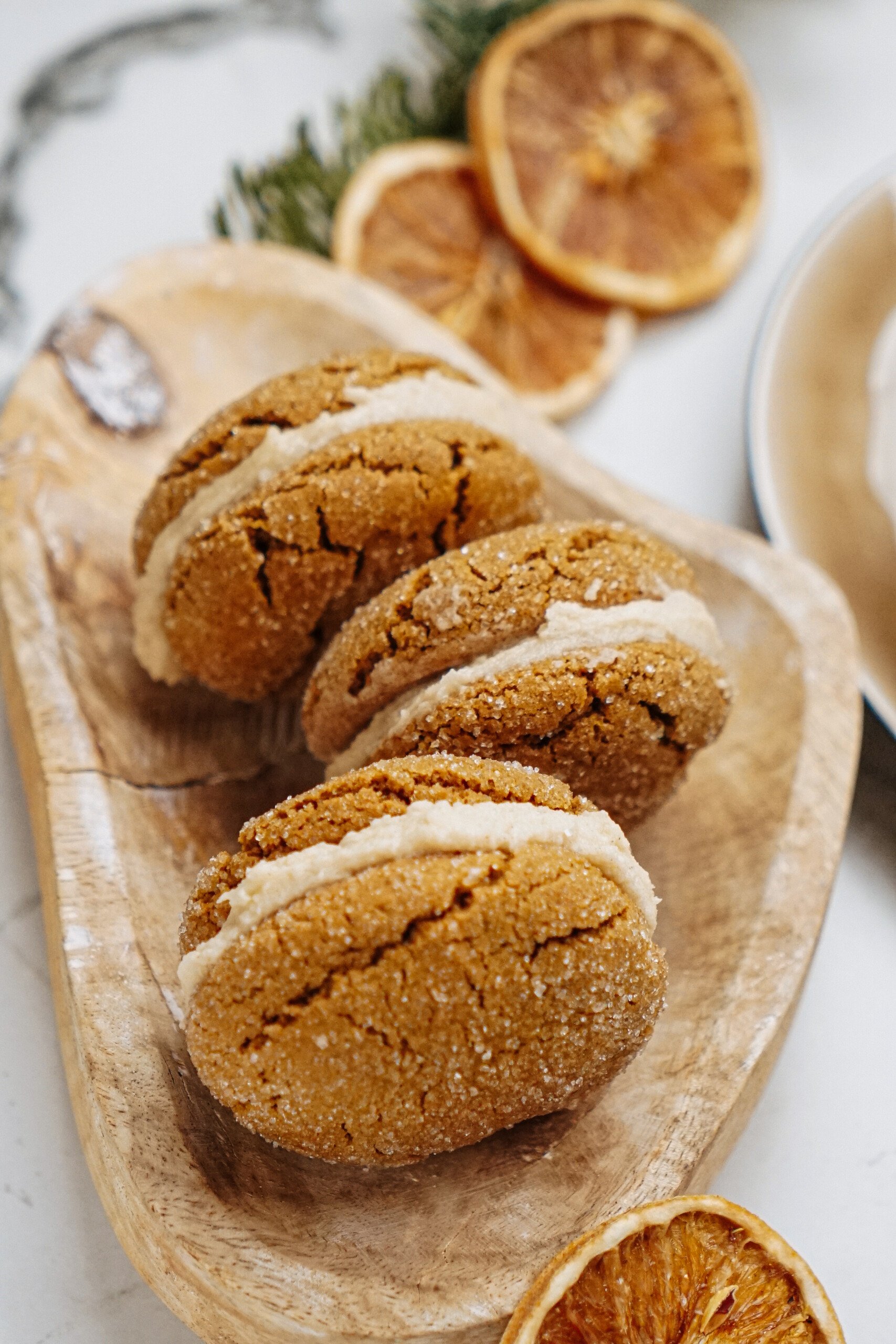 Three pumpkin whoopie pies with creamy filling sit on a wooden tray, accompanied by decorative dried orange slices and adding an extra layer of indulgence.