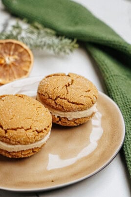 Two ginger cookies with cream filling rest on a beige plate, accompanied by a green cloth and a dried orange slice in the background, reminiscent of hazelnut shortbread's rich, nutty charm.