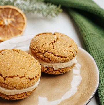 Two ginger cookies with cream filling rest on a beige plate, accompanied by a green cloth and a dried orange slice in the background, reminiscent of hazelnut shortbread's rich, nutty charm.