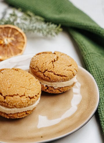 Two ginger cookies with cream filling rest on a beige plate, accompanied by a green cloth and a dried orange slice in the background, reminiscent of hazelnut shortbread's rich, nutty charm.