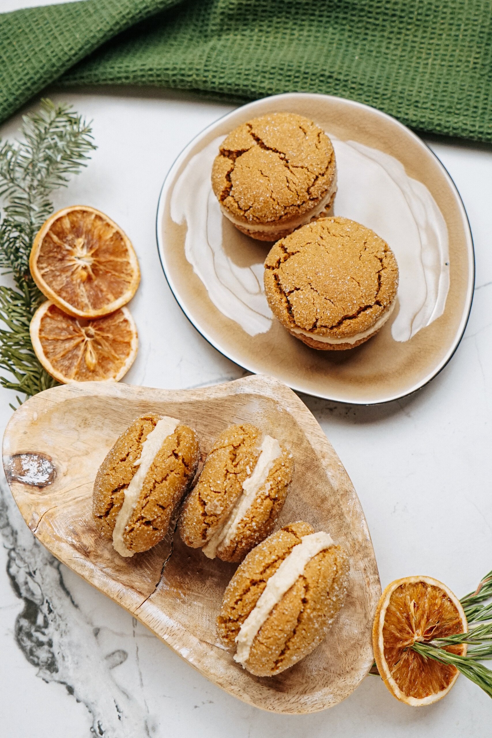 gingerdoodle cookies filled with cream rest on two plates, surrounded by dried orange slices and greenery on a marble surface.