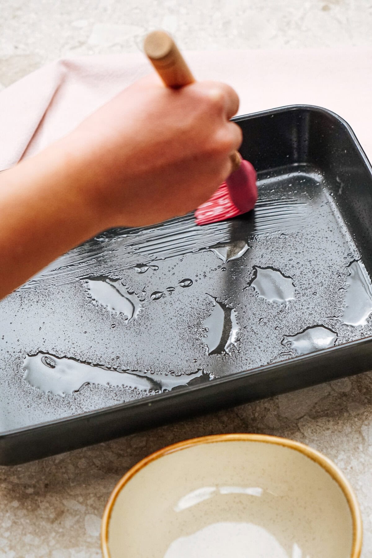 A hand using a pink brush to spread oil on a black baking tray, with a beige bowl nearby.