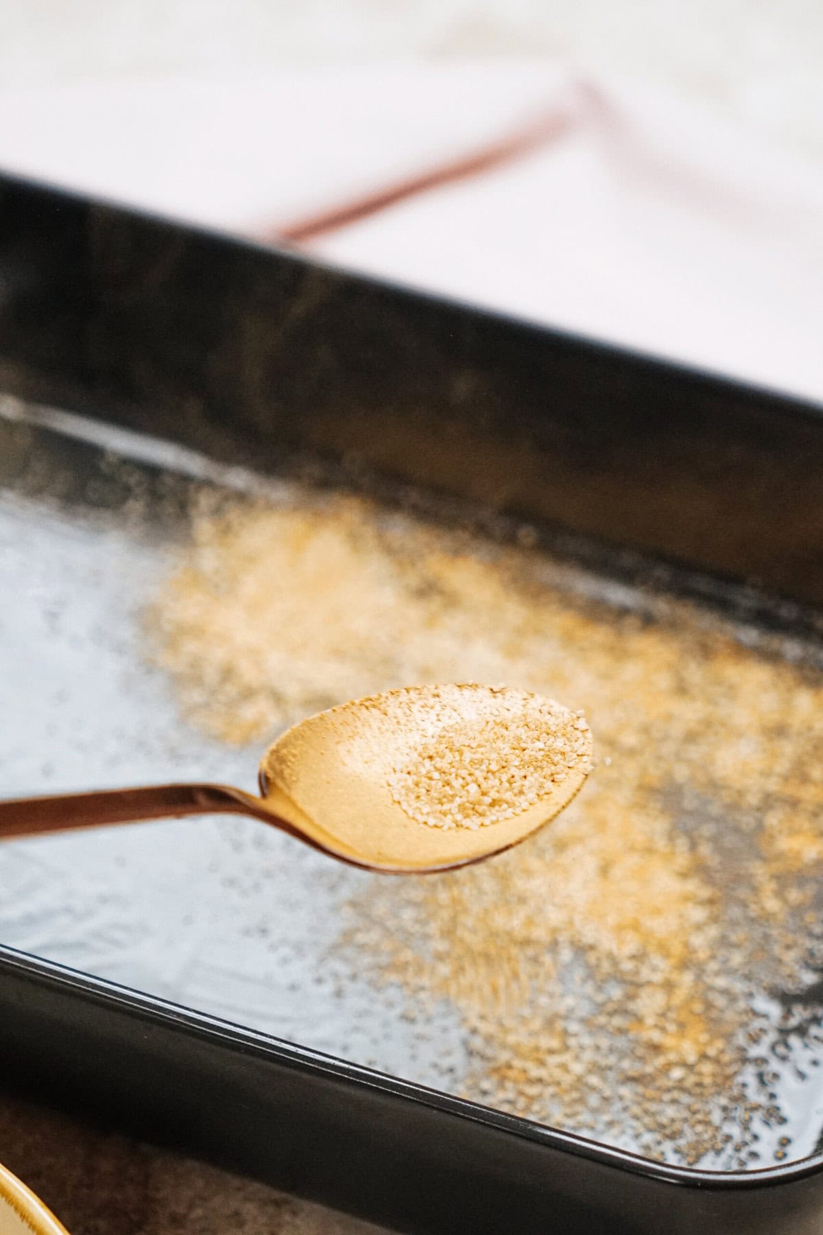 A spoon holds a small amount of granulated brown sugar over a black baking tray.