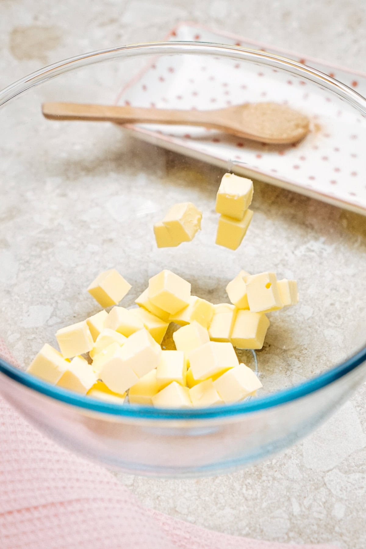 Cubes of butter in a glass bowl on a marble countertop, with a small wooden spoon resting on a polka dot plate in the background.