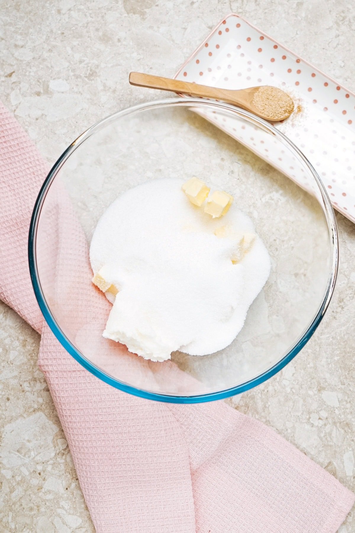 A mixing bowl with sugar and butter sits on a marble surface next to a pink cloth and a small plate with a spoonful of sugar.