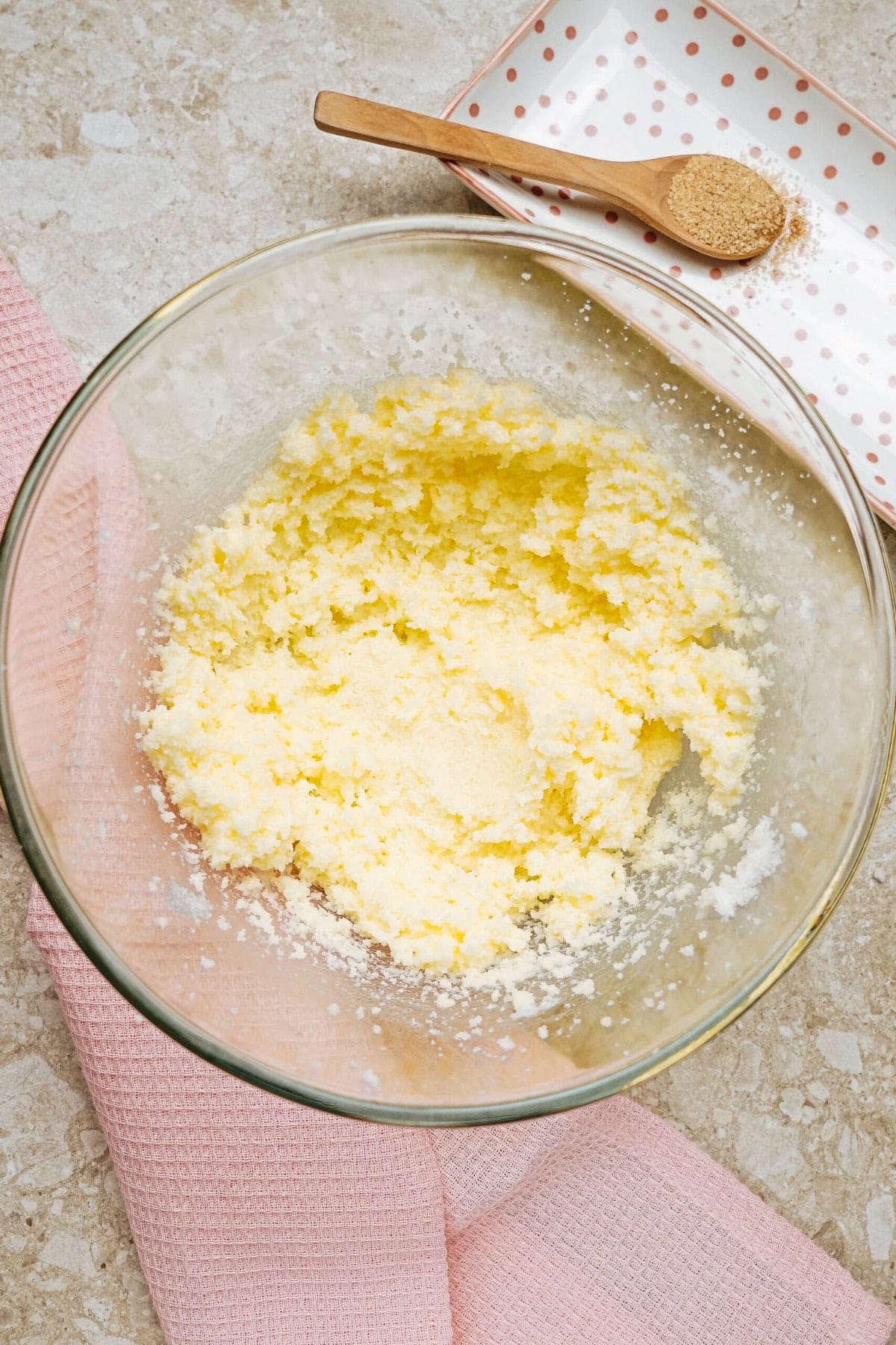 A glass bowl with creamed butter and sugar on a countertop, accompanied by a pink cloth and a wooden spoon on a polka dot plate.