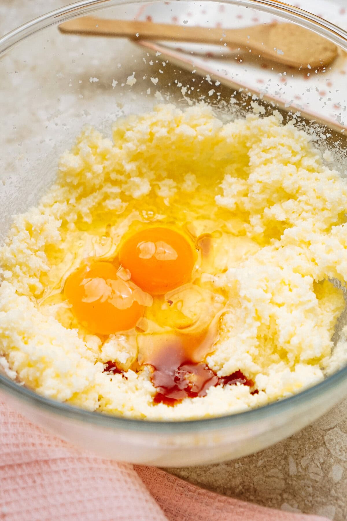 A glass bowl with three cracked eggs and vanilla extract atop creamed sugar and butter. A pink cloth and wooden spoon are nearby.