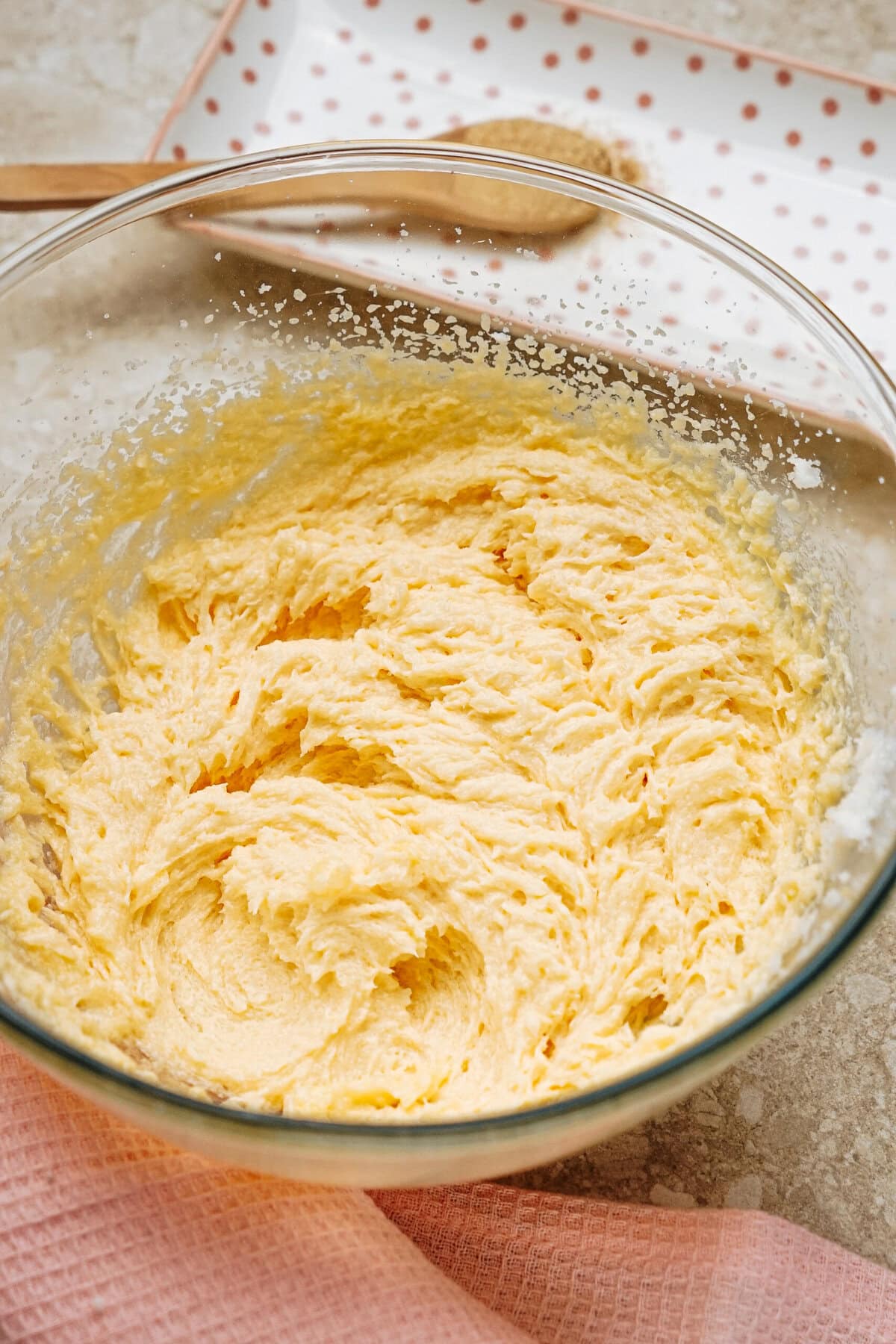 A glass bowl of creamy, yellow cake batter on a kitchen countertop, with a wooden spoon and a polka dot plate in the background.