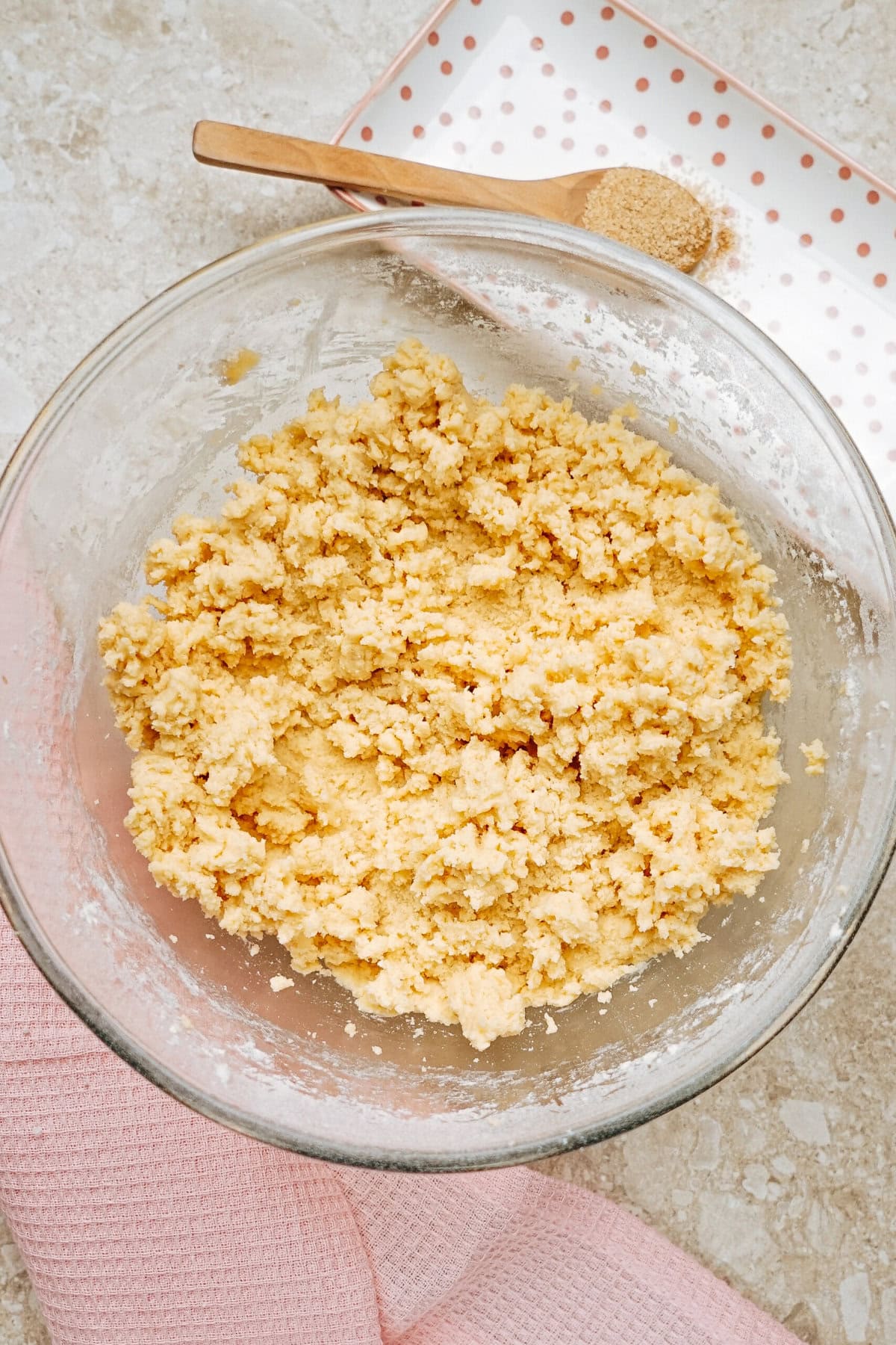 A glass bowl filled with crumbled dough mixture sits on a countertop. A wooden spoon with brown sugar is on a polka dot tray in the background, next to a pink cloth.