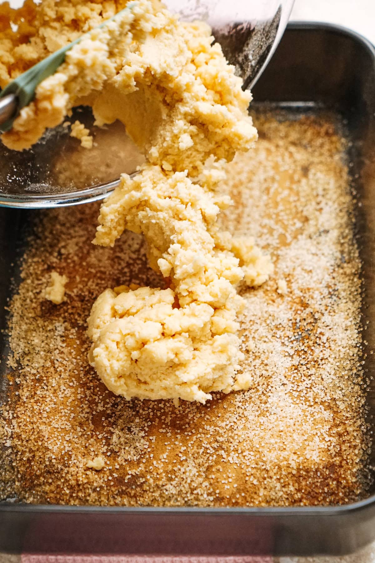 Thick, yellow cake batter being poured from a bowl into a deep, greased baking pan dusted with breadcrumbs.