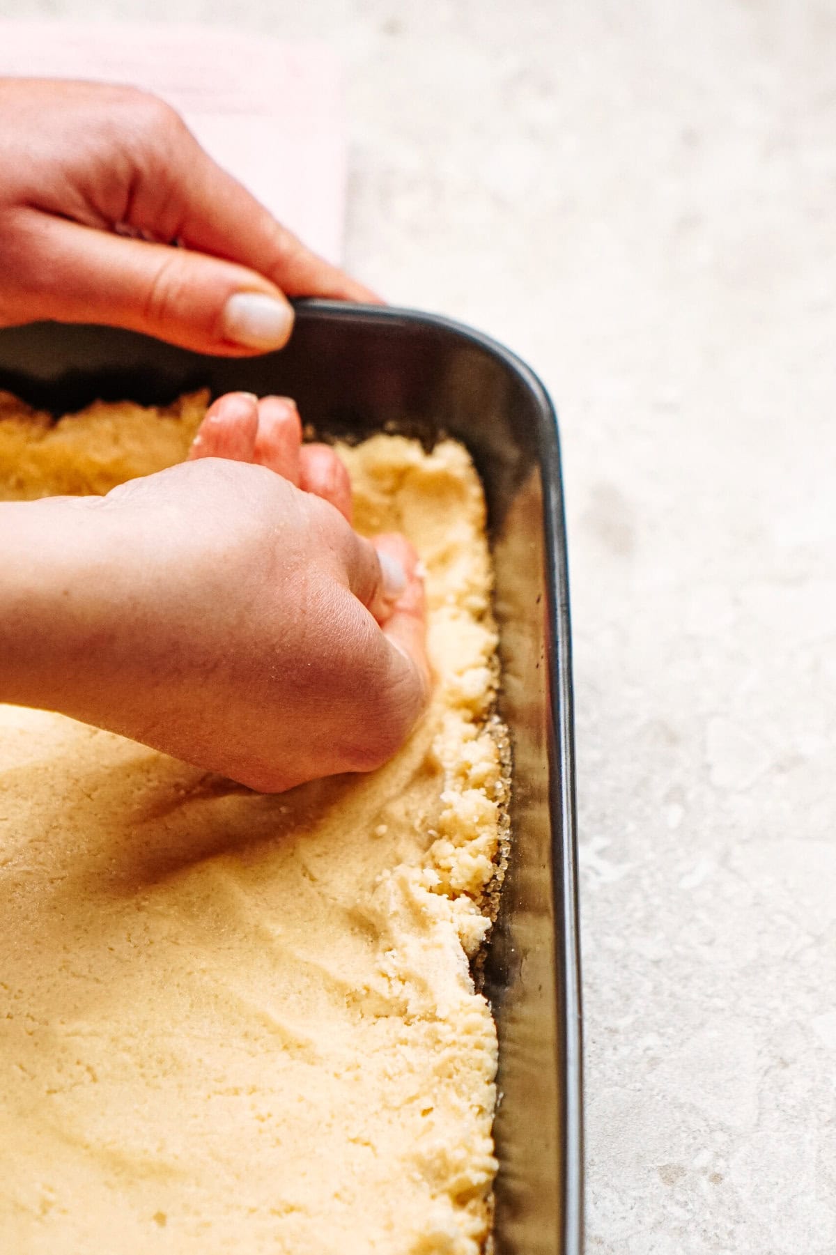 Hands pressing dough into a rectangular baking pan on a light-colored countertop.