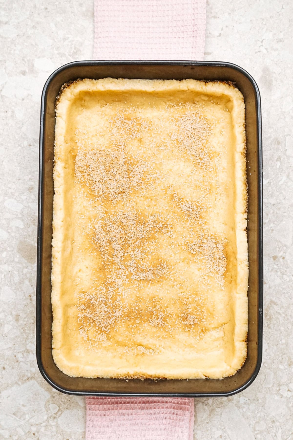 A rectangular baking dish with a freshly baked cakelike dessert dusted with sugar on a textured surface.