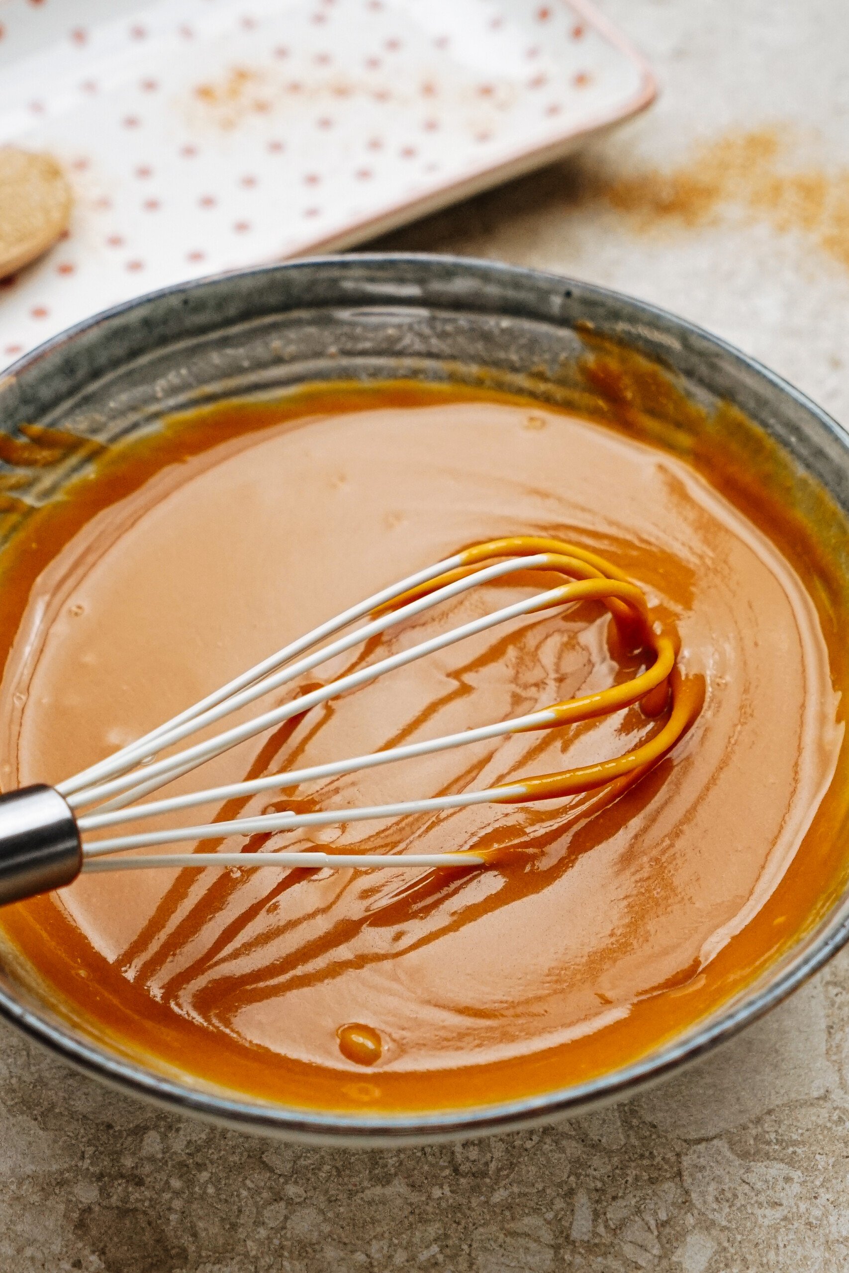 A whisk in a bowl of smooth caramel sauce, with a small, polka-dotted plate in the background.